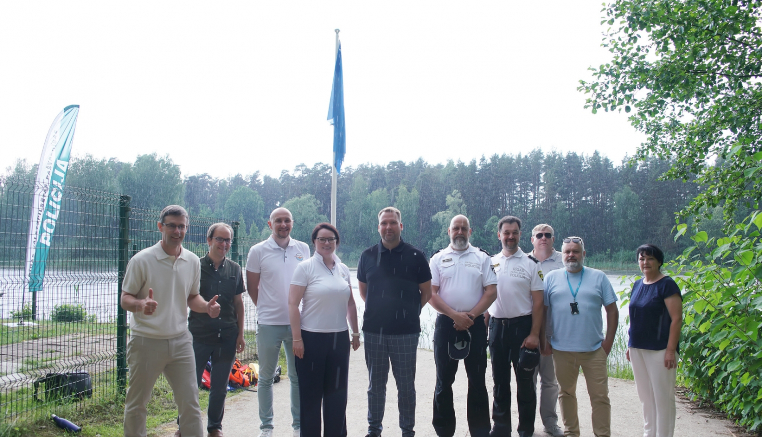 Cilvēki fotografējas pie Bābelīša ezra zilā karoga