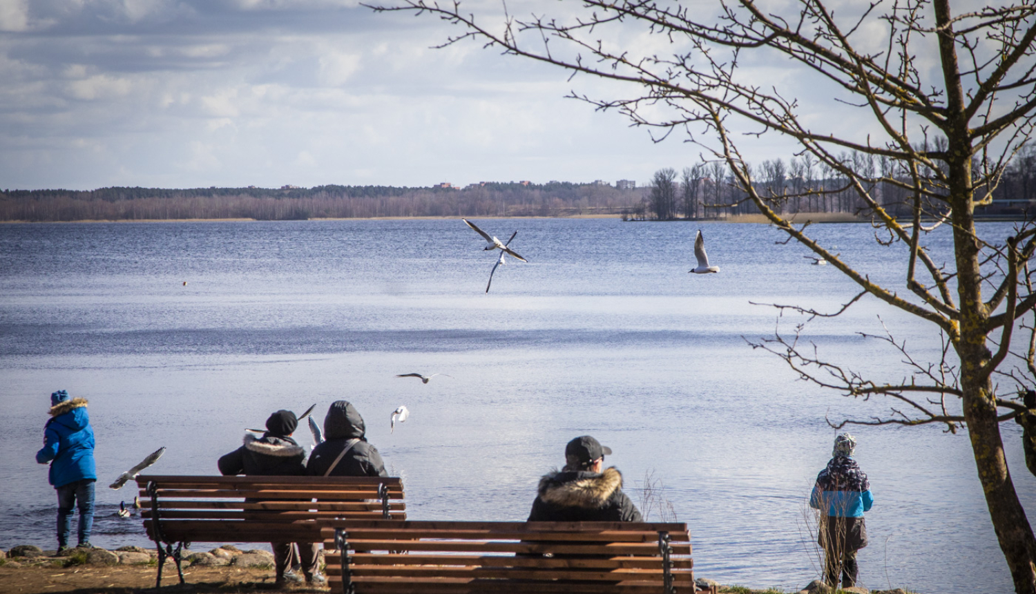 Cilvēki sēž uz soliņiem ezera krastā