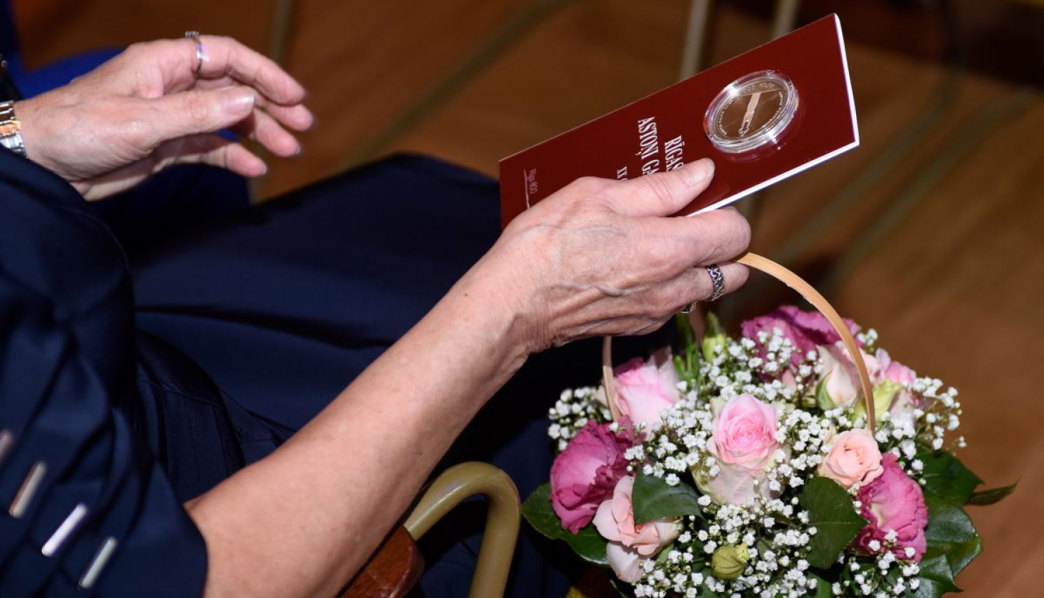 Rokās tur ziedus un jubilejas sudraba monētu