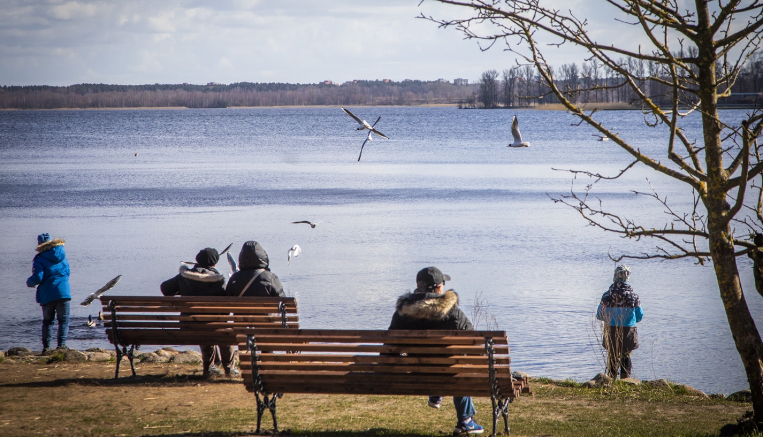 Cilvēki sēž uz soliņiem ezera krastā